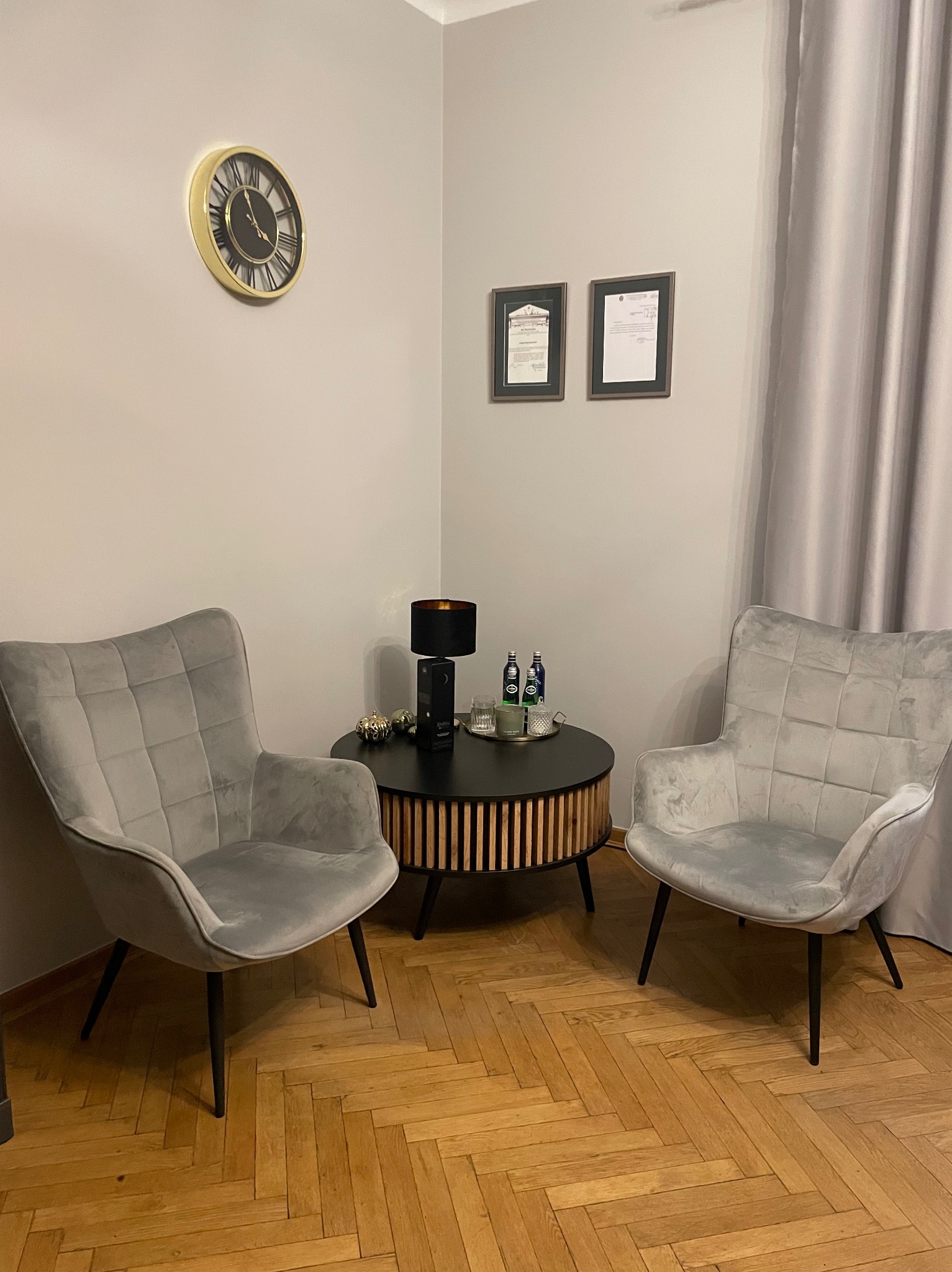 Wnętrze biura z dwoma szarymi fotelami, okrągłym stolikiem z napojami i lampą, dyplomami na ścianie i zegarem.