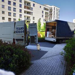 Przetransportowani Cezary Stefańczuk - Dwa busy transportowe, jeden z otwartymi drzwiami, zaparkowane na brukowanym podjeździe przed nowoczesnym budynkiem mieszkalnym, z widoczną platformą na kółkach.