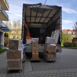 Przetransportowani Cezary Stefańczuk - Częściowo załadowana ciężarówka z otwartą paką, z widocznymi kartonami zabezpieczonymi folią stretch i taśmami transportowymi, stojąca na brukowanym podjeździe przed budynkiem.