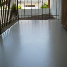 Gładka, jasnoszara powierzchnia tarasu balkonu z metalową balustradą i fragmentem drewnianych drzwi, widok na budynek w tle.