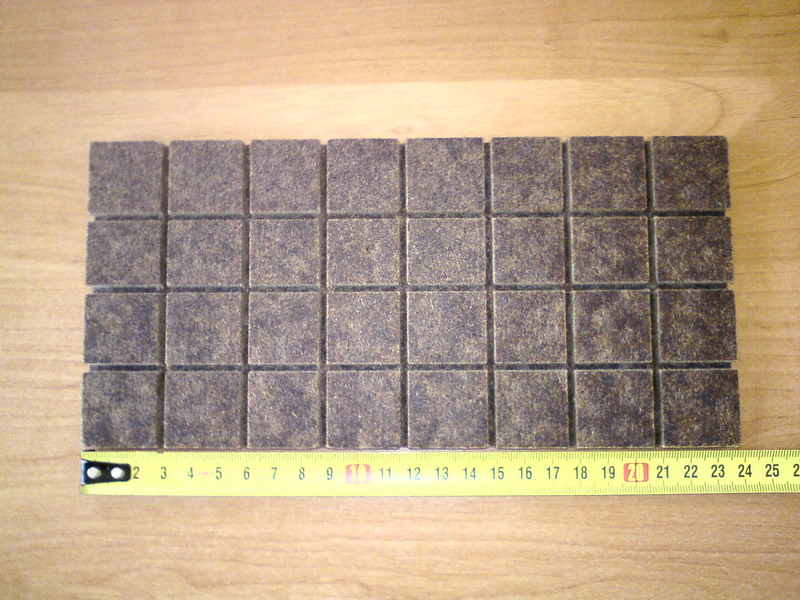 Kwadratowe brykiety z płyty pilśniowej miękkiej ułożone obok żółtej miarki na drewnianym blacie. Przybliżenie na strukturę materiału.