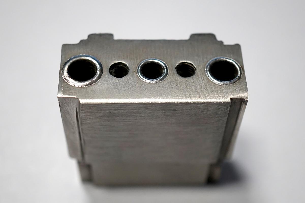 Metalowy blok z precyzyjnie wywierconymi otworami o różnej średnicy, widoczne ślady obróbki skrawaniem.
