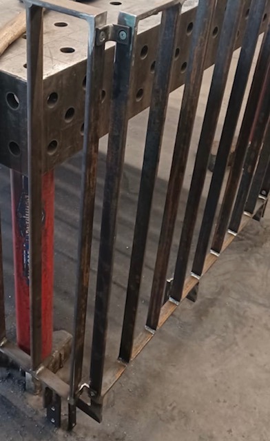 Metalowa konstrukcja z pionowych prętów, spawana do ramy na stole spawalniczym, widoczne ślady spawania i niedoskonałości powierzchni.