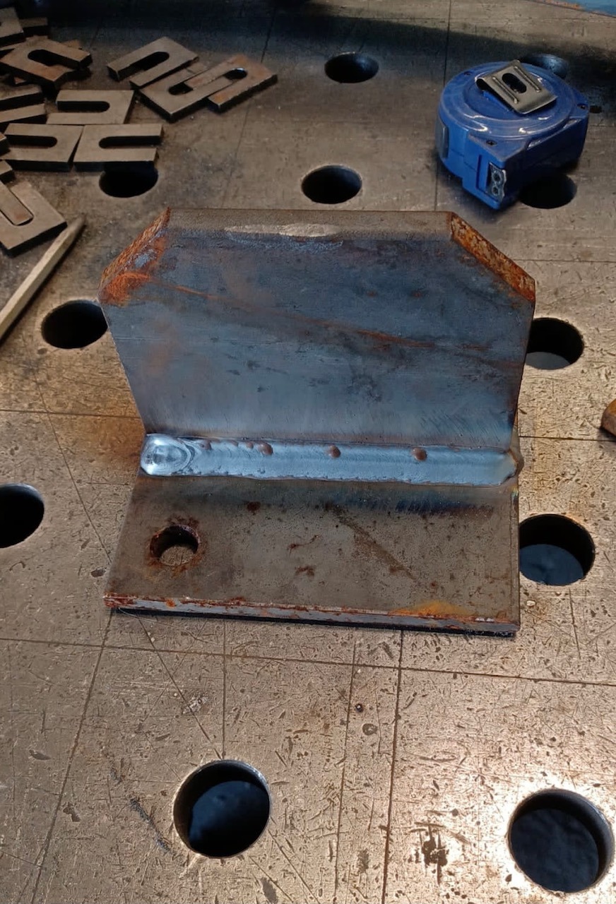 Połączenie spawane dwóch elementów stalowych, widoczna spoina i ślady rdzy na krawędziach, na perforowanym stole spawalniczym, obok miara zwijana i metalowe podkładki.