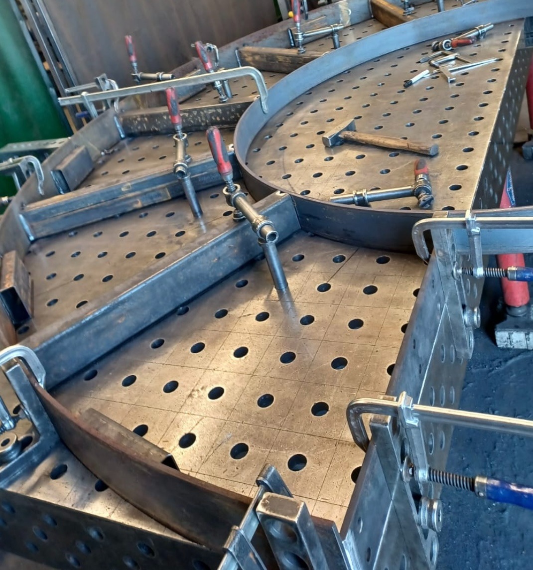 Metalowa konstrukcja z profili mocowana do stołu spawalniczego za pomocą ścisków, widoczny młotek ślusarski.