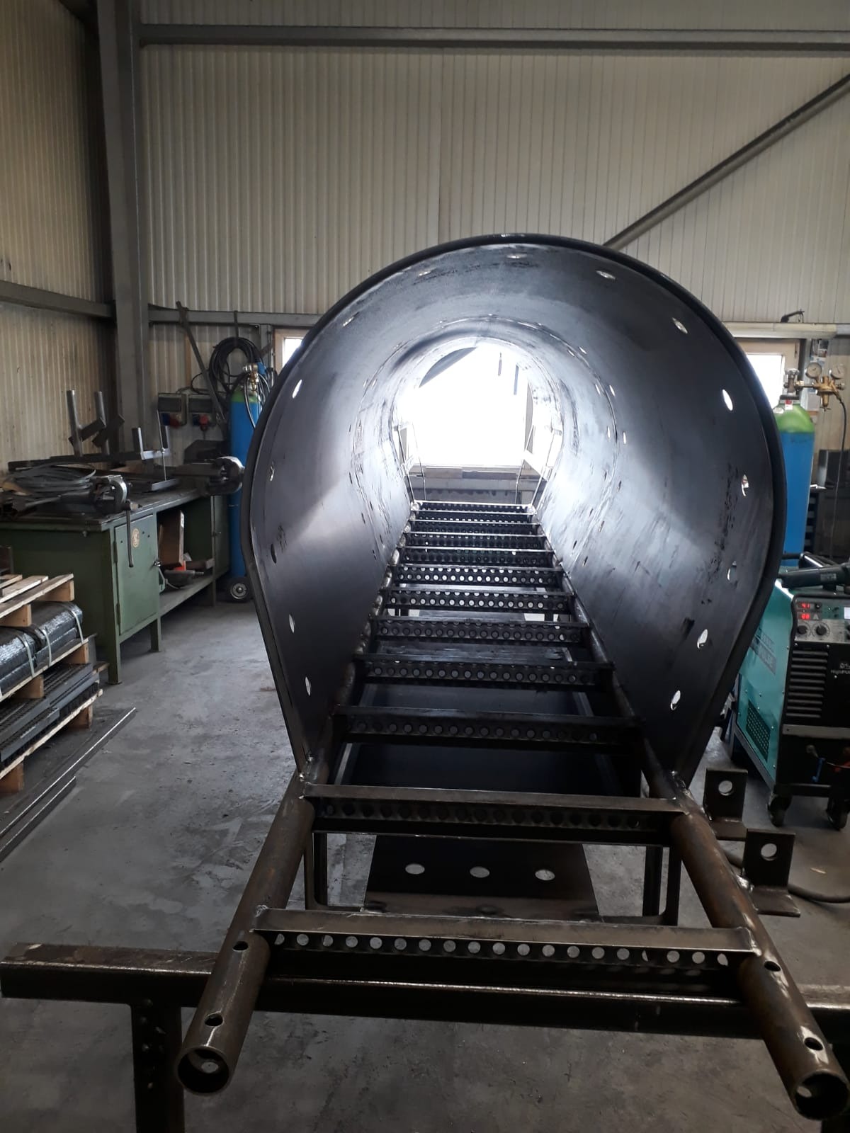 Metalowa konstrukcja z perforowanymi stopniami wewnątrz cylindrycznej obudowy, widok od dołu w warsztacie.