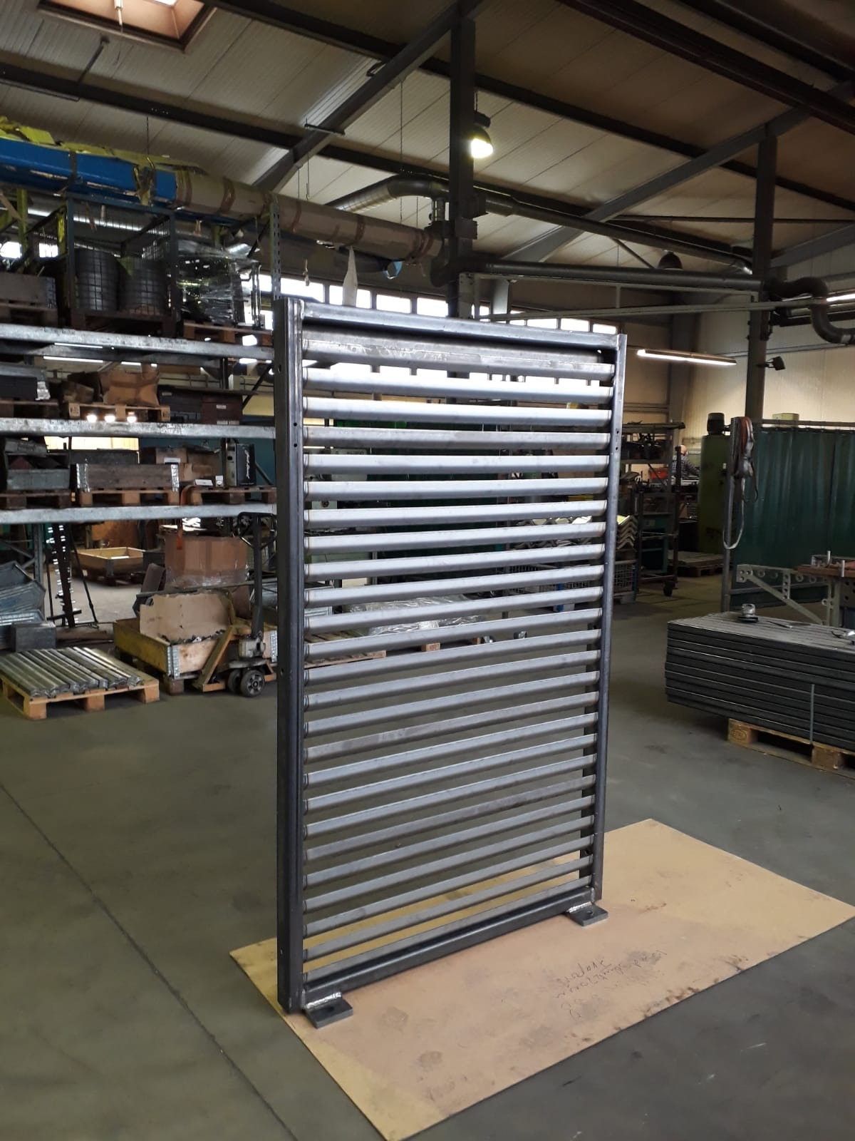 Metalowa, spawana konstrukcja z poziomych profili, stojąca na kartonowej podkładce w hali produkcyjnej.