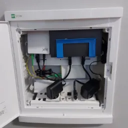 Otwarta biała szafka telekomunikacyjna marki EPN Elegant z zainstalowanym routerem KDX, zasilaczami i kablami Ethernet, światłowodowymi oraz zasilającymi, zamontowana na szarej ścianie.