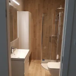 Nowoczesna łazienka z kabiną prysznicową, umywalką z szafką i toaletą, wykończona płytkami imitującymi drewno, z widoczną listwą LED nad lustrem.