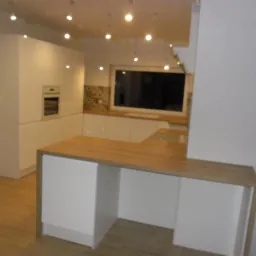 Nowoczesna, biała kuchnia z drewnianym blatem, oświetleniem punktowym i oknem z widokiem na zewnątrz, w trakcie wykańczania, widoczne ślady po pracach remontowych.