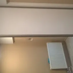 Biała szafa przesuwna z szarymi obramowaniami, odbicie wnętrza pokoju z grzejnikiem i panelami podłogowymi w lustrzanych drzwiach.