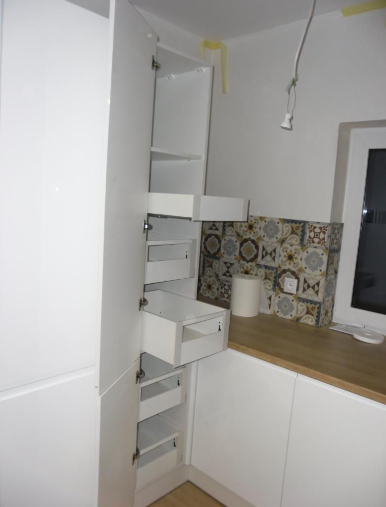 Biała szafka kuchenna z otwartymi drzwiami i wysuwanymi szufladami, widoczne wnętrze z półkami, tło stanowią płytki dekoracyjne i okno.