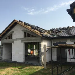 Budowa domu z białych bloczków, częściowo pokryty ciemną dachówką, widoczne rusztowania i przygotowane pakiety dachówek na dachu, błękitne niebo w tle.