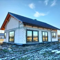 Surowy stan domu murowanego z ciemną dachówką w Andrychowie, widoczne okna z czarnymi ramami i częściowo ośnieżony teren wokół.