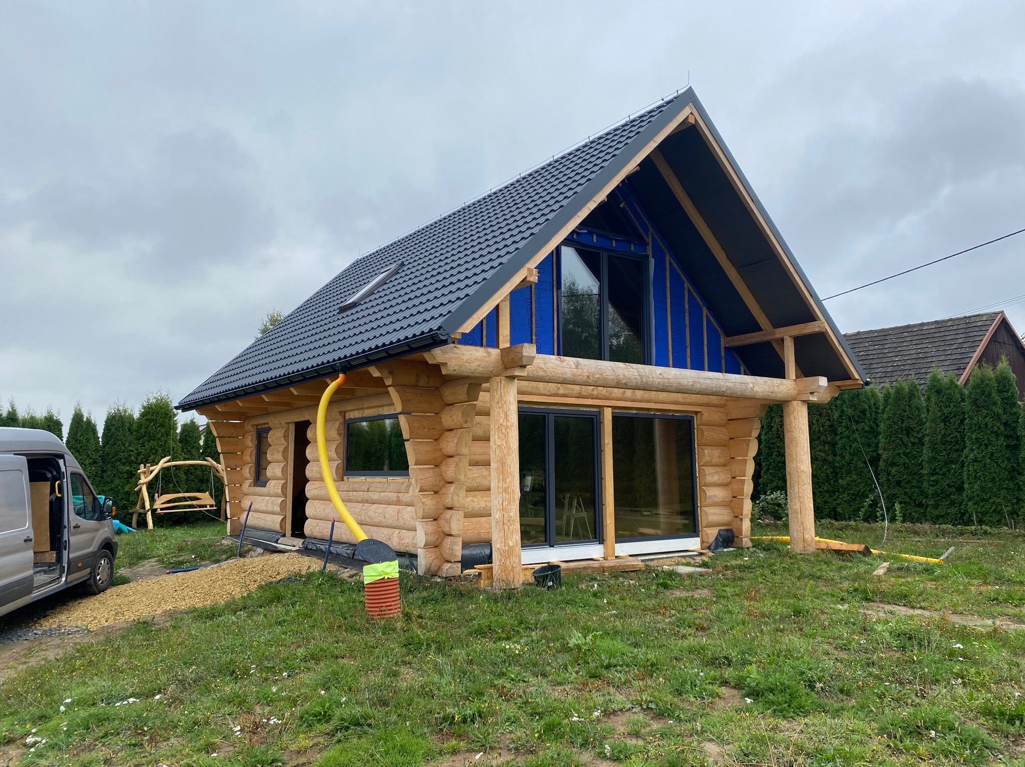 Surowy drewniany dom z ciemnym dachem w trakcie budowy, widoczne okna bez wykończenia, rury drenażowe i częściowo zagospodarowany teren.