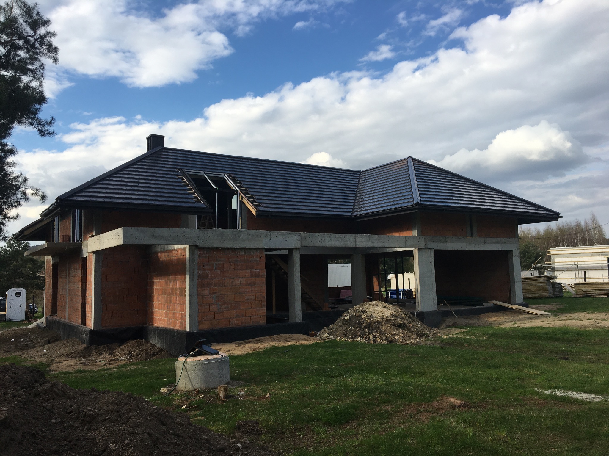 Surowy stan domu murowanego z ciemnym dachem, widoczne cegły, betonowe filary i otwarte okna dachowe, na zewnątrz plac budowy z ziemią i trawą pod błękitnym niebem.