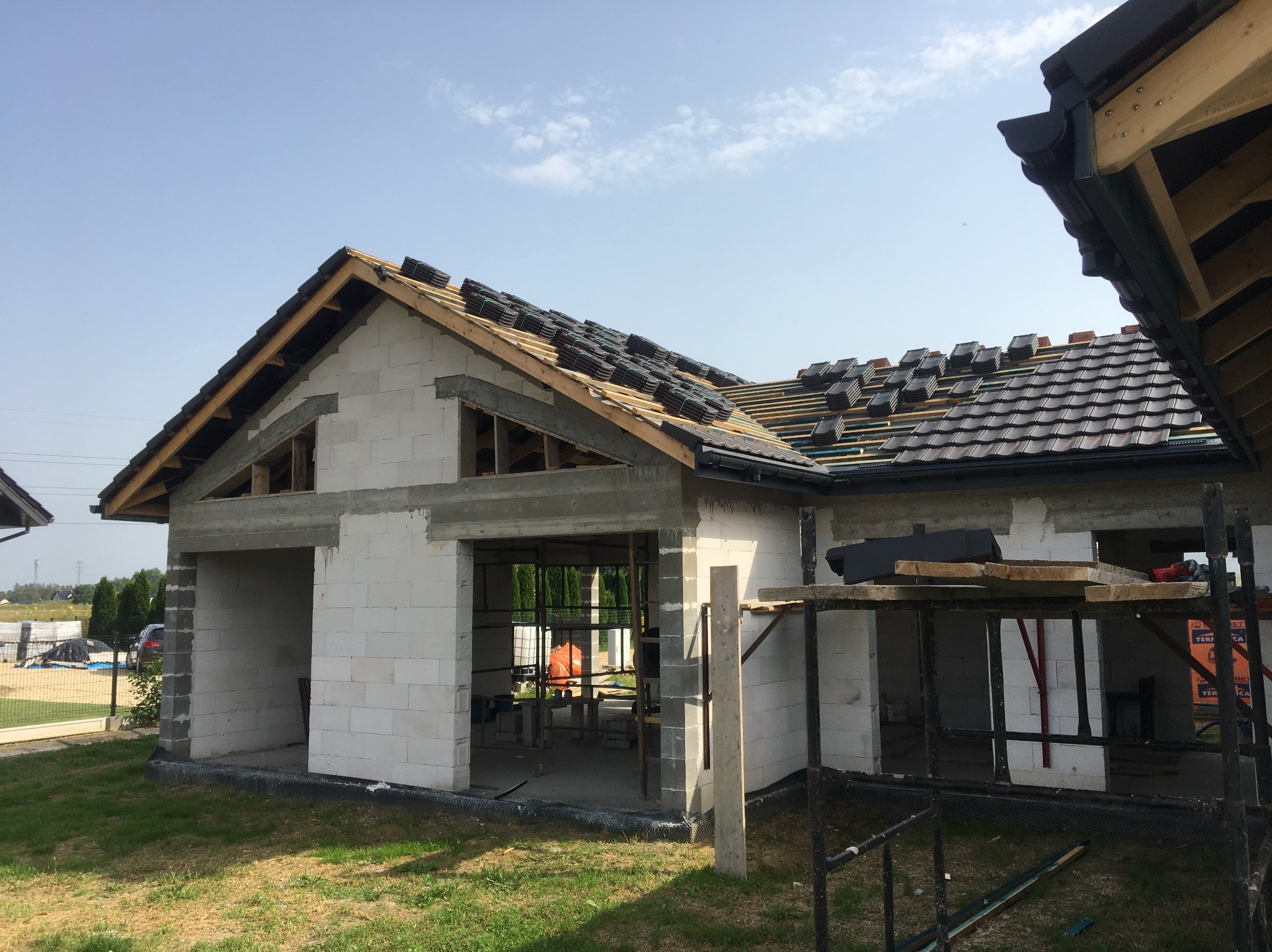 Budowa domu z białych bloczków, częściowo pokryty ciemną dachówką, widoczne rusztowania i przygotowane pakiety dachówek na dachu, błękitne niebo w tle.