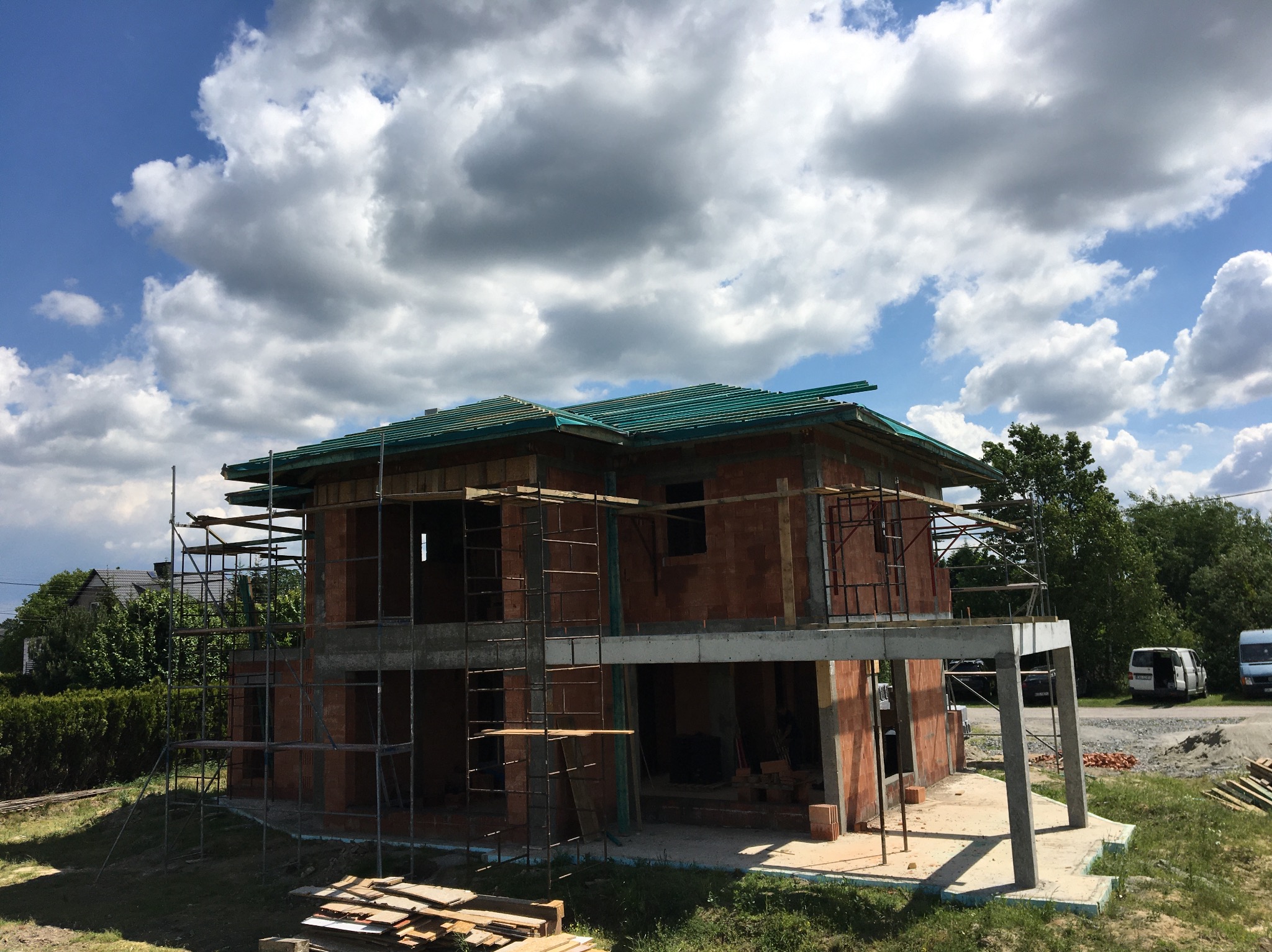 Surowy, dwukondygnacyjny budynek z czerwonej cegły w trakcie budowy, z widocznymi rusztowaniami i wstępnie ułożonymi elementami dachu; w tle błękitne niebo z białymi chmurami.
