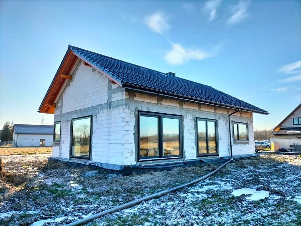 Surowy stan domu murowanego z ciemną dachówką w Andrychowie, widoczne okna z czarnymi ramami i częściowo ośnieżony teren wokół.