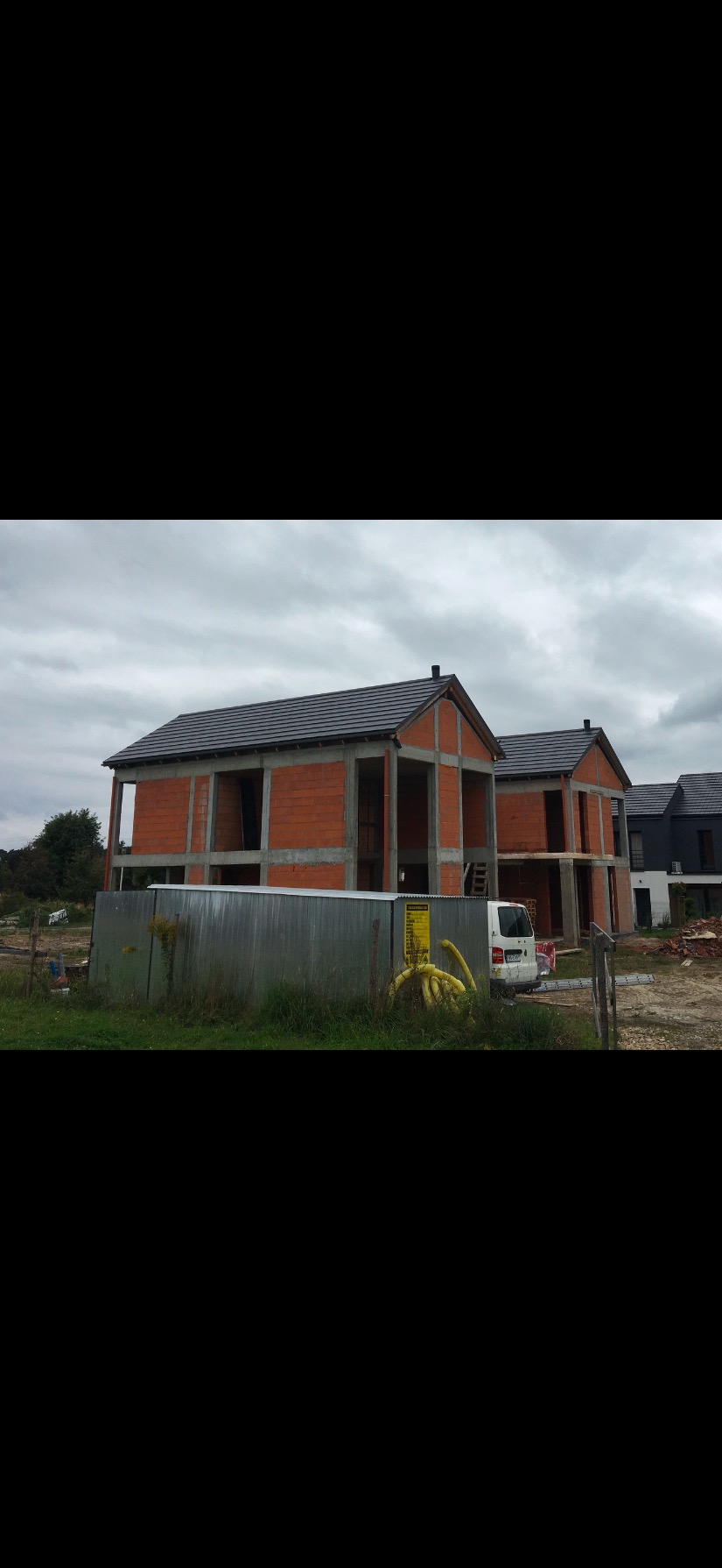 Stan surowy dwóch domów murowanych z ciemną dachówką w Andrychowie, widok zza blaszanej bariery i zaparkowanego białego busa.