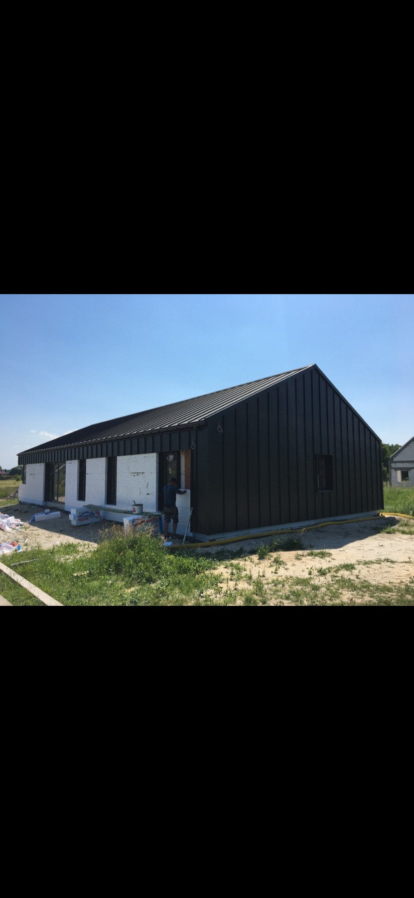 Nowoczesny dom jednorodzinny z ciemną blachą na elewacji i dachu, w trakcie montażu ocieplenia okien, widoczne materiały izolacyjne i pracownik.