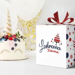 Tort jednorożec z owocami i białe pudełko prezentowe z logo 'Lukrowa Kraina' na białym tle z balonami.