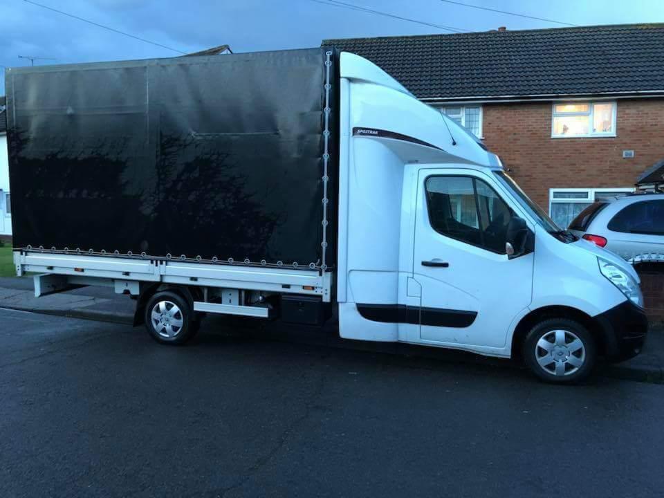 Biała furgonetka z czarną plandeką zaparkowana na ulicy przed domem z cegły.