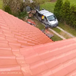 Czerwona dachówka ceramiczna widziana z góry, pracownik w kamizelce odblaskowej przycina dachówkę na przenośnym stole warsztatowym obok srebrnego vana z otwartymi drzwiami, narzędzia i materiały...