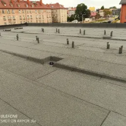 Szary, płaski dach z papy termozgrzewalnej z widocznymi wpustami dachowymi i uchwytami do instalacji, na tle zabudowy miejskiej.