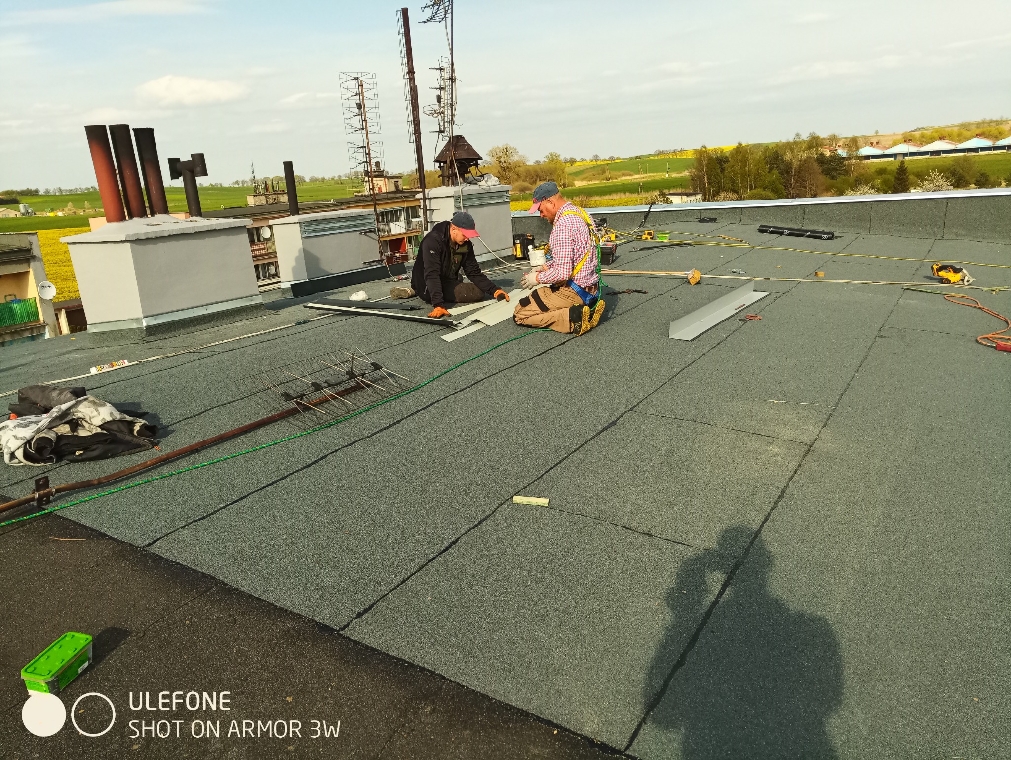 Dwóch dekarzy w uprzężach, klęczących na płaskim dachu pokrytym papą, dopasowuje metalowe elementy obróbek blacharskich; w tle kominy, anteny i pola uprawne.