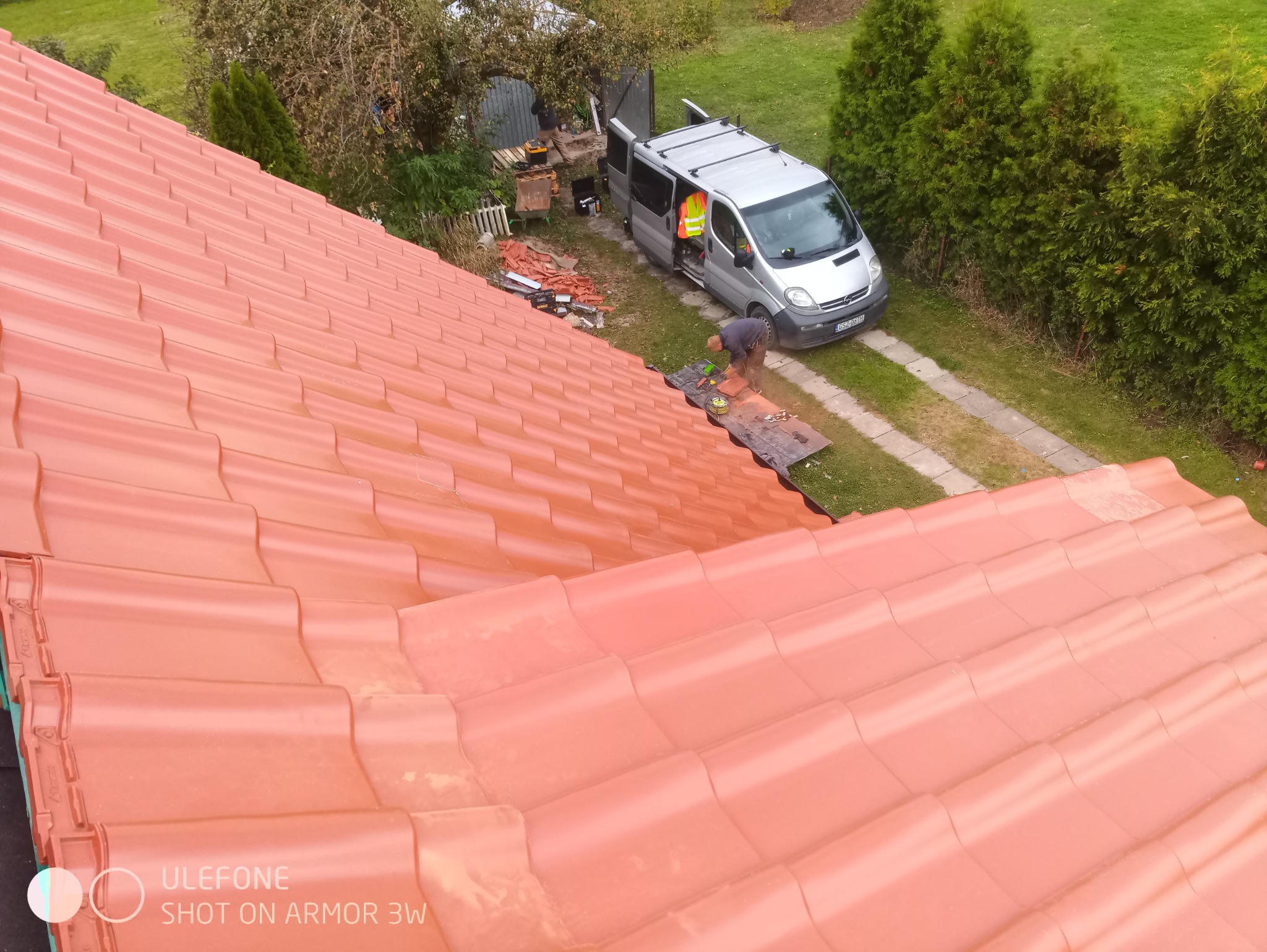 Czerwona dachówka ceramiczna widziana z góry, pracownik w kamizelce odblaskowej przycina dachówkę na przenośnym stole warsztatowym obok srebrnego vana z otwartymi drzwiami, narzędzia i materiały...