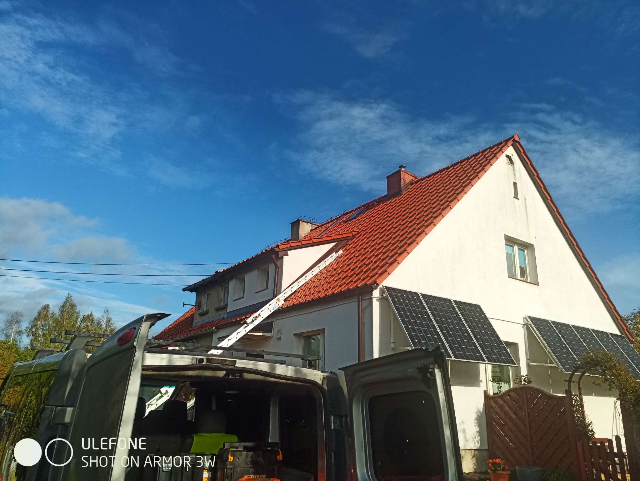 Dom jednorodzinny z czerwonym dachem, drabina oparta o dach i panele słoneczne na elewacji, widok z wnętrza samochodu dostawczego.