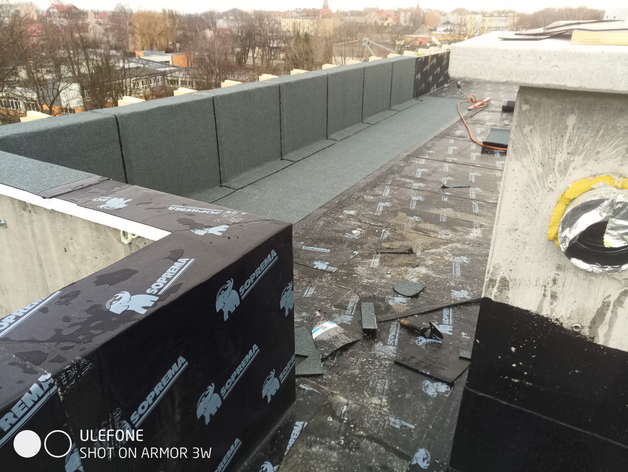 Widok z góry na płaski dach w trakcie krycia papą termozgrzewalną marki Soprema, widoczne narzędzia i elementy izolacji, w tle zabudowa miejska.