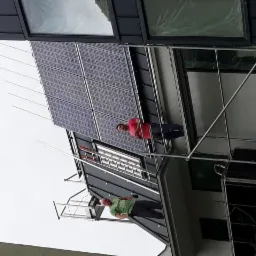 Dwóch monterów na rusztowaniu instaluje panele słoneczne na dachu domu z czarną blachodachówką. Widoczne zabezpieczenia przeciw ptakom. Jeden monter w zielonej koszulce, drugi w czerwonej.