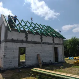 Wznoszenie więźby dachowej z zielonych belek na budynku z białych bloczków, dwóch pracowników na dachu, w tle błękitne niebo z chmurami i drzewa.