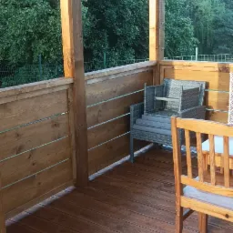 Zadaszony taras z drewnianą podłogą i meblami ogrodowymi. Widok na zieleń w tle. Ciepłe światło podkreśla teksturę drewna.