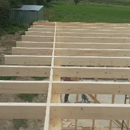 Wysoki kąt ujęcia na drewniany szkielet podłogi w trakcie budowy, z zielonym krajobrazem w tle i widocznym fragmentem narzędzi.