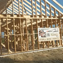 Konstrukcja szkieletowa domu w trakcie budowy, widoczna więźba dachowa i drewniane elementy nośne. Na pierwszym planie baner reklamowy firmy. Słoneczny dzień.