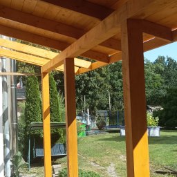 DOM-DREW Budowa domów tradycyjnych murowanych i szkieletowych drewnianych. - Drewniana konstrukcja z belek, prawdopodobnie zadaszenie tarasu. Widoczne słupy nośne i elementy dachu na tle zieleni ogrodu. Jasne drewno, słoneczny dzień.