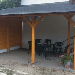 DOM-DREW Budowa domów tradycyjnych murowanych i szkieletowych drewnianych. - Zadaszona altana z drewnianymi słupami i ścianką, stół z krzesłami wewnątrz. Dach kryty ciemną dachówką bitumiczną. Brukowana podłoga, doniczka z kwiatami.