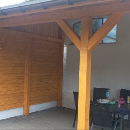 DOM-DREW Budowa domów tradycyjnych murowanych i szkieletowych drewnianych. - Drewniana konstrukcja z zadaszeniem i ścianką z desek, wsparta na solidnym słupie. W tle okno w białej ścianie. Na zewnątrz stoi stół z krzesłami.