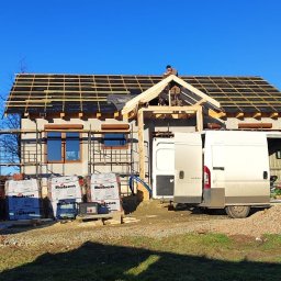 DOM-DREW Budowa domów tradycyjnych murowanych i szkieletowych drewnianych. - Budowa domu szkieletowego w trakcie: widoczna konstrukcja dachu z łatami, okna z roletami, rusztowania, palety z dachówkami Roben i dwa busy dostawcze na tle błękitnego nieba.