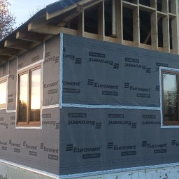 DOM-DREW Budowa domów tradycyjnych murowanych i szkieletowych drewnianych. - Budowa domu szkieletowego: widok na ścianę z oknami, pokrytą membraną Eurovent, z widoczną konstrukcją więźby dachowej i pracującym mężczyzną w tle.