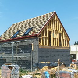 DOM-DREW Budowa domów tradycyjnych murowanych i szkieletowych drewnianych. - Rozpoczęta budowa domu z drewnianą konstrukcją dachu z łatami i dwoma oknami dachowymi, szkieletem poddasza oraz czarną membraną na parterze, otoczona rusztowaniem.