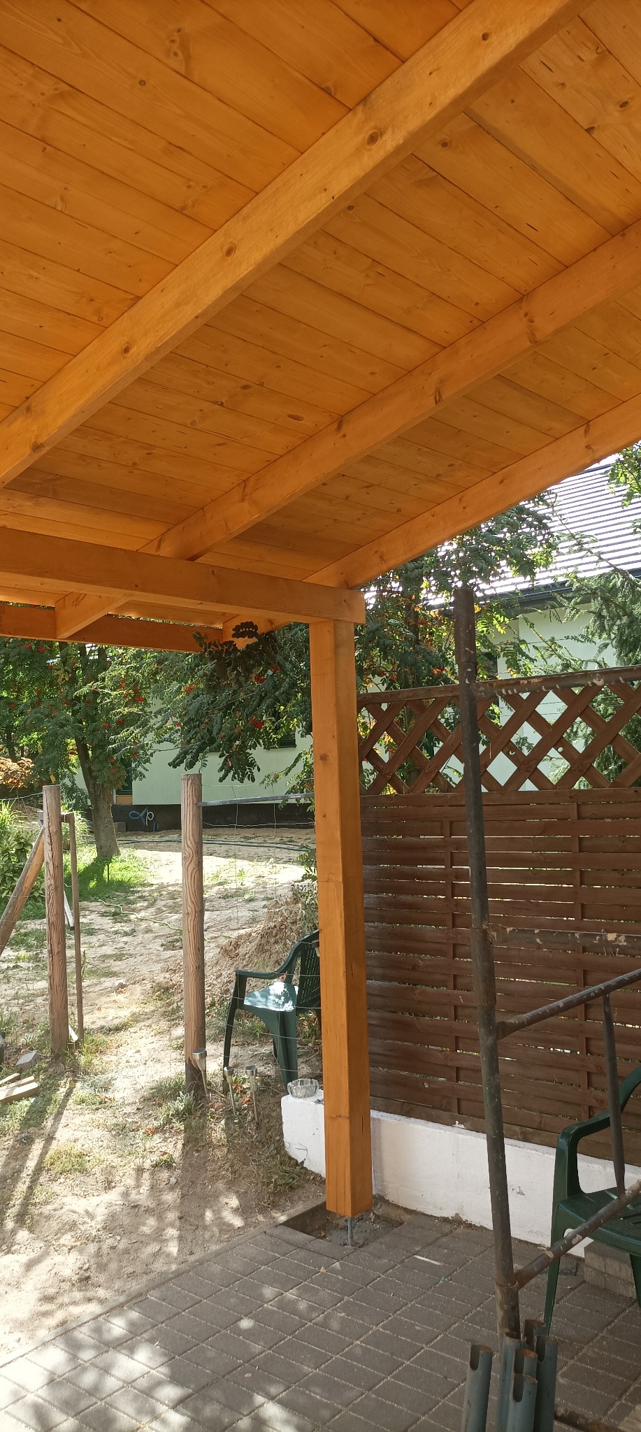 Drewniana konstrukcja z belek, prawdopodobnie zadaszenie lub altana, w trakcie budowy, widoczne elementy mocujące i otoczenie ogrodowe.