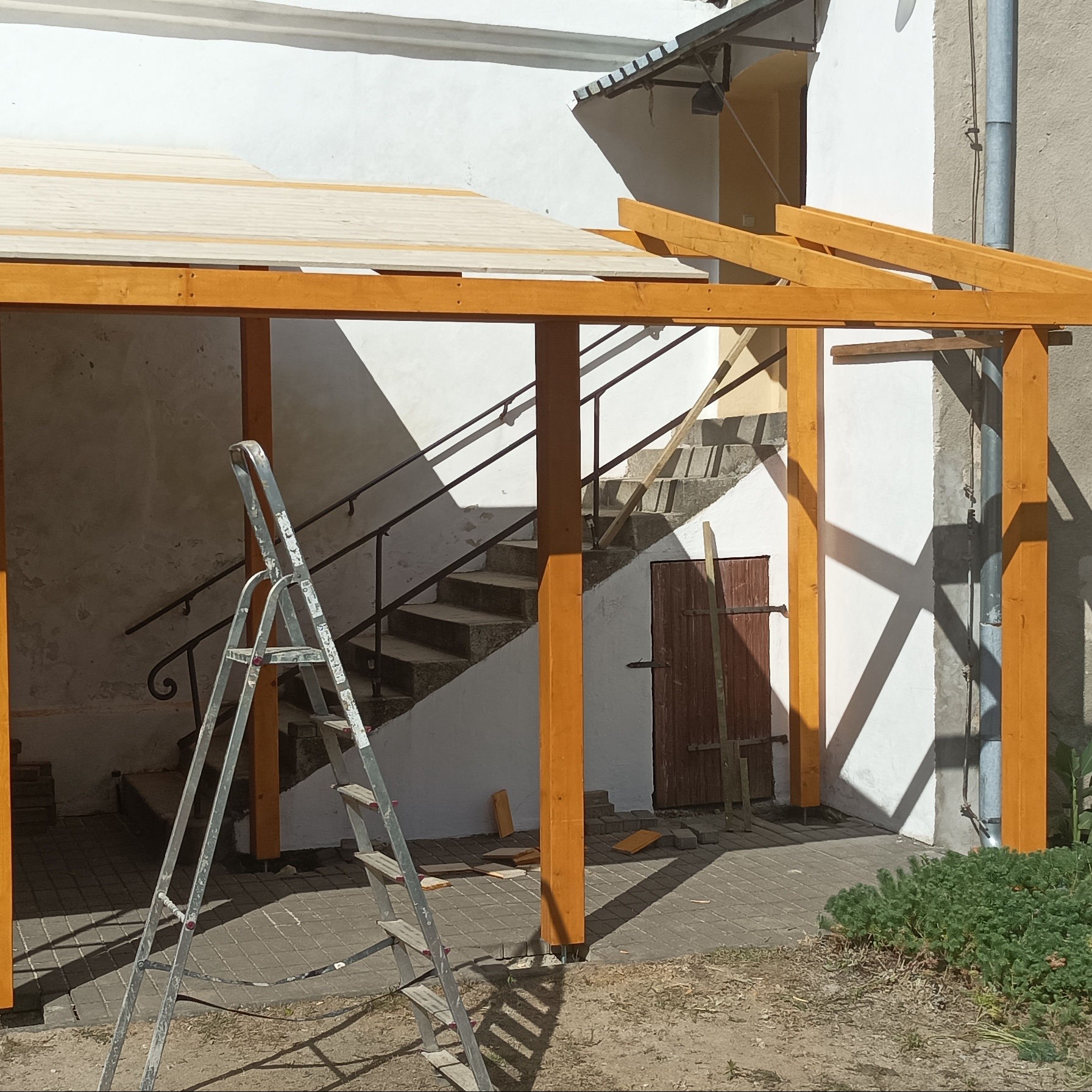 Konstrukcja z belek w ciepłym odcieniu drewna, częściowo zadaszona, w trakcie budowy. Aluminiowa drabina oparta o konstrukcję. W tle schody i budynek.