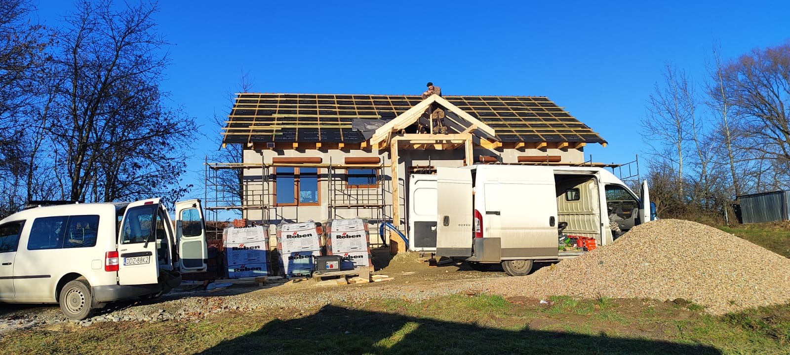 Budowa domu szkieletowego w trakcie: widoczna konstrukcja dachu z łatami, okna z roletami, rusztowania, palety z dachówkami Roben i dwa busy dostawcze na tle błękitnego nieba.