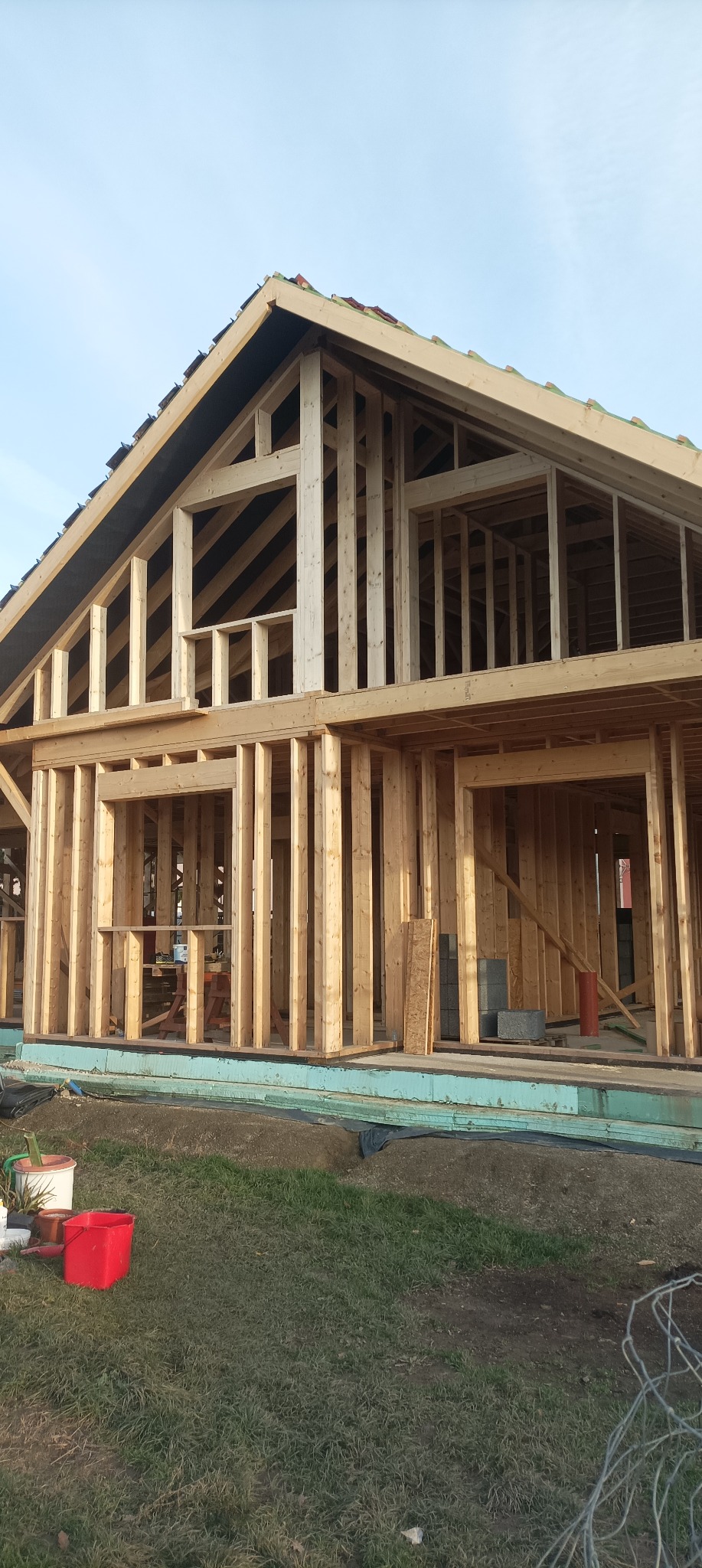 Surowy stan domu szkieletowego w budowie, widoczne drewniane ramy ścian i dachu, z elementami izolacji i materiałów budowlanych na zewnątrz. Ujęcie z dołu.