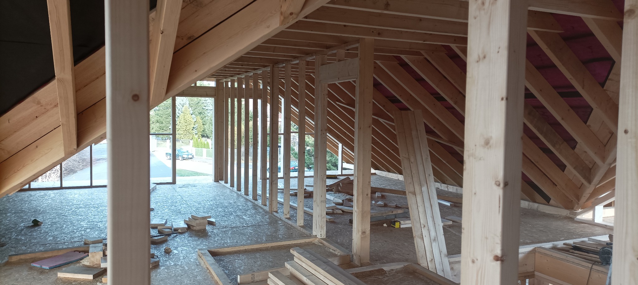 Wnętrze domu w trakcie budowy szkieletowej. Widoczna konstrukcja drewniana ścian, dachu i podłogi. Na zewnątrz okien zieleń drzew i zabudowa.
