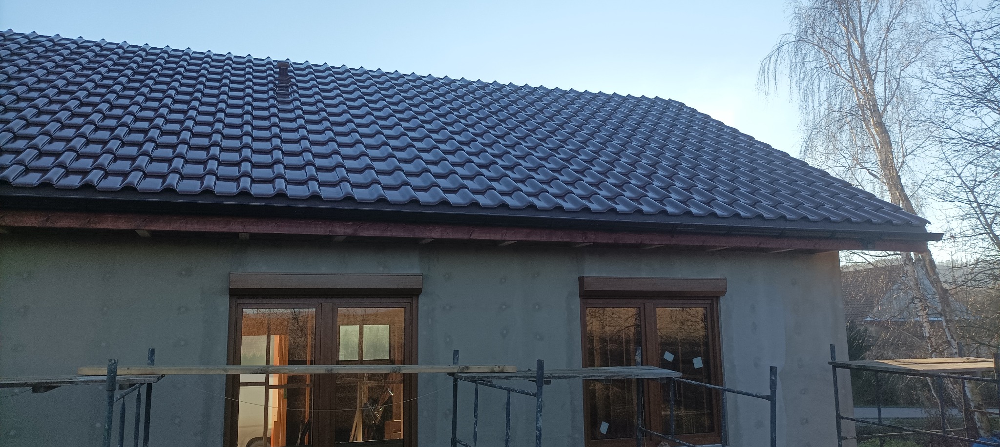 Ciemna dachówka ceramiczna na nowym domu z szarym tynkiem, widoczne rusztowania i okna z roletami. Ujęcie z dołu, błękitne niebo w tle.